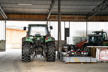 Modern green good-looking tractor stands in outside garage near agricultural fixtures and transport on farm or ranch on winter or summer cloudy day. Agriculture industry, cargo, bio production, 