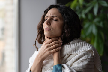 Fototapeta na wymiar Upset sick woman touching painful swollen neck glands, suffering from throat infection, unhealthy female standing near window at home feeling pain with swallowing, selective focus