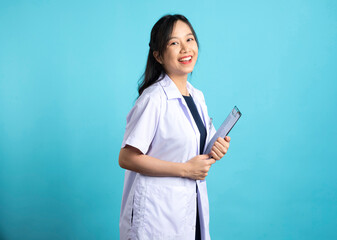 Asian doctor woman in medical uniform and clipboard smiling at camera standing on blue white background.