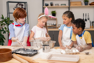 Children pouring dough together, wearing aprons looking at each other with wide smiles while preparing appetizing cookies for their parents