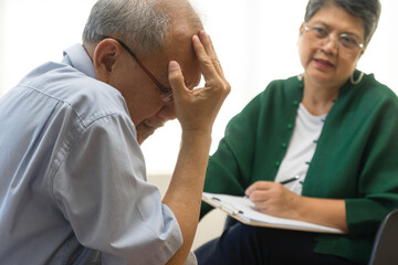 Fototapeta premium Depressed Asian senior man sharing problems during therapy session with female psychiatrist writing on clipboard.