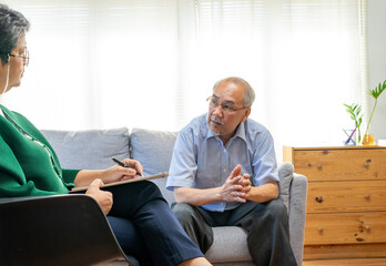 Depressed Asian senior man sharing problems during therapy session with female psychiatrist writing on clipboard. - obrazy, fototapety, plakaty