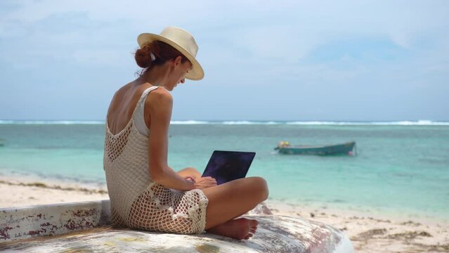 Successful business woman in yellow hat working on laptop by the sea. Pretty lady typing on computer at summer day outdoors. Freelance, travel and holidays concept.