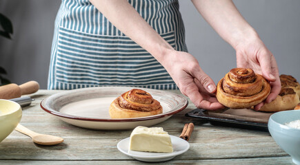 Woman in a blue apron is taking out fresh warm cinnamon rolls buns from a baking tray and putting them on a plate. Concept of delicious homemade pastries and cozy atmosphere