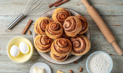 There is a plate with homemade fresh delicious cinnamon rolls, ingredients for their preparation and kitchen tools on the wooden table . Top view