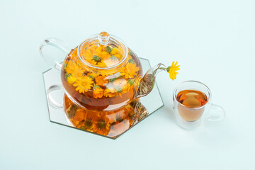 Herbal tea. Transparent glass teapot, cups of tea with orange flowers of calendula on a blue background. Calming drink concept - obrazy, fototapety, plakaty