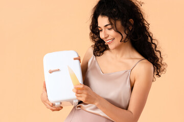 Young smiling woman holding refrigerator and bottle of cosmetic product on color background