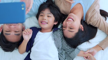 Happy family time, Taking selfie mobile phone of Asian parents dad, mom and son laying together on bed in bedroom with happy, laughing smiles. Weekend parenting with love good time relaxing at home.