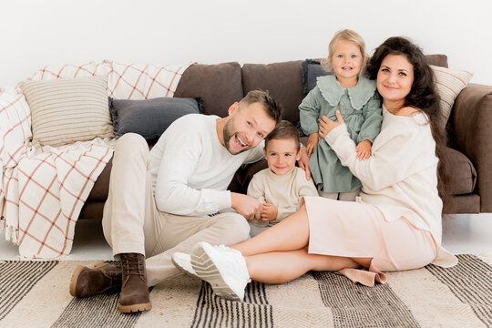 Happy beautiful parents sitting near the sofa with joyful little kids, smiling, spending weekends at home. Charming father with pretty mother playing with cute daughter and son, family concept
