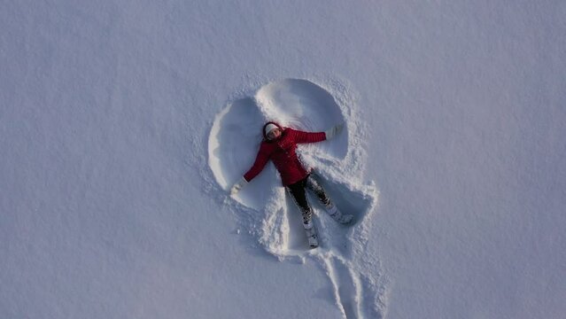 Aerial view from drone of girl in red winter coat making snow angel. Girl waves arms and legs lying in snow. High quality 4k footage