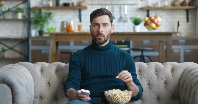 Shocked bearded man watching horror movie on TV and eating popcorn while sitting on the couch at home. Scared millennial male watching terrible news on TV channel while sitting on sofa at living room