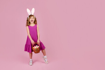Full body little beautiful smiling girl in Easter bunny ears holds a basket with eggs on pink...
