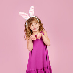 Obraz na płótnie Canvas Adorable funny little kid with curly blond hair in dress and bunny ears, smiling and showing paws gesture while imitating rabbit during Easter party in pink studio