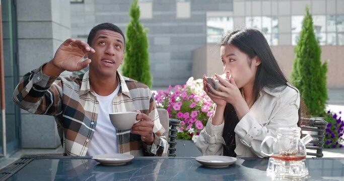 Charming multi-ethnic couple in love sits at a table on the summer terrace of a cozy cafe, holding cups of tea in hands, talking with pleasure to each other during a date on a beautiful warm sunny day