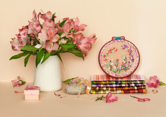 Spring still life with a bouquet of pink alstroemeria flowers, hand embroidery with satin ribbons in hoop and a set of multi-colored embroidery ribbons on a pale pink pastel background. DIY concept