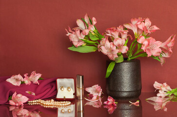 Romantic still life with delicate pink alstroemeria flowers in a vase, pearl beads and earrings and lipstick on a burgundy background with reflection. Festive mood, date, gifts concept