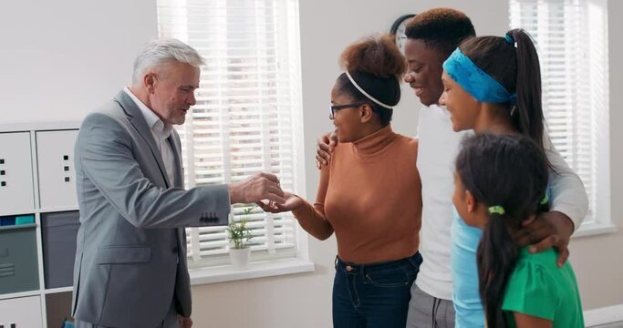 An elderly man with gray hair, a real estate agent sells an apartment to a young couple with children, hands the woman the keys explaining the details, the family hugs with happiness
