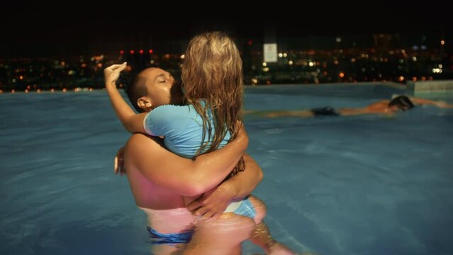 The young love couple enjoy each other in roof illumination pool in hotel. Laughing man twist in whirl in embrace cute woman in water pool. Night city view. Concept positive smile laughter emotions.