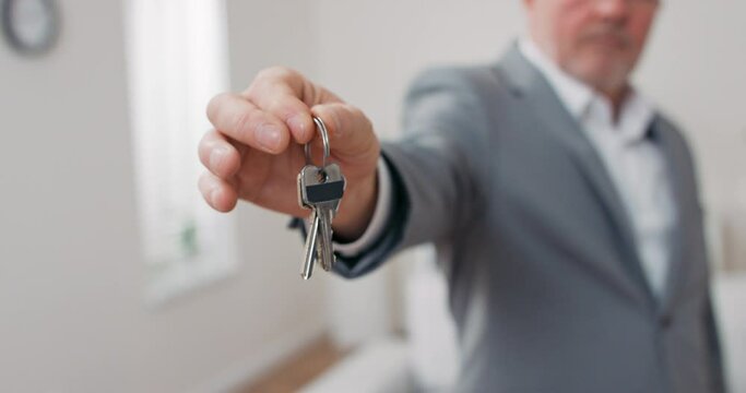 Shot of apartment keys extended on palm of hand toward camera, gray-haired mature man dressed in suit hands over keys, real estate agent, owner of apartment, gives room to use