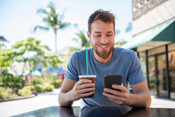 Phone app young man using technology device holding cellphone at cafe texting with messaging sms apps drinking coffee in summer. Handsome young casual man using smartphone smiling happy. Urban male. - Powered by Adobe