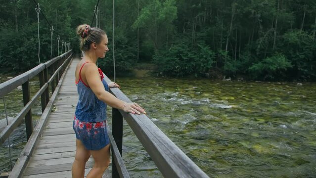 Woman Walking Along Suspension Bridge Alone in Picturesque Green Forest Setting. Slow Motion of Dark Haired Woman Walk on Suspension Bridge in Thick Green Forest. No Other People Are in The Shot.