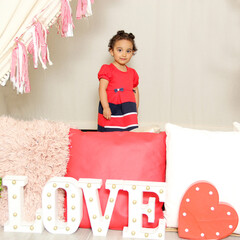 Latin little girl with wavy hair 2 years old in red dress under a happy teepee with a sign that...