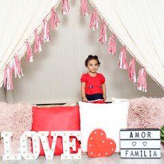 Latin little girl with wavy hair 2 years old in red dress under a happy teepee with a sign that...