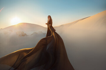 Mystery arabic woman in black long dress stands in desert long train silk fabric fly flytter in wind motion. clothes gold accessories hide face. Oriental fashion model. Sand dunes background sunset. - obrazy, fototapety, plakaty