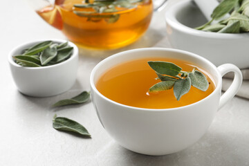 Cup of sage tea and green leaves on white table