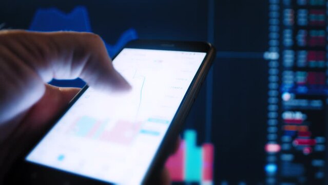 Person hand holds a smartphone with a stock market app and analyze data against the background of a defocused graph close-up