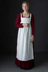 A Regency maid servant wearing a red linen dress with an apron and a cap against a studio backdrop