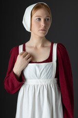 A Regency maid servant wearing a red linen dress with an apron and a cap against a studio backdrop - obrazy, fototapety, plakaty