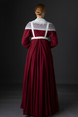 A Regency maid servant or working woman wearing a red linen dress with an apron and a lace modesty shawl against a studio backdrop