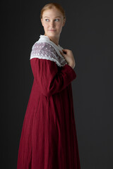 A red-haired Regency woman wearing a simple red linen dress with a lace modesty shawl against a studio backdrop