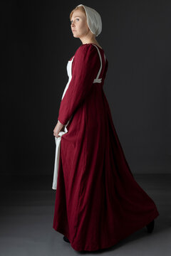 A Regency Maid Servant Wearing A Red Linen Dress With An Apron And A Cap Against A Studio Backdrop