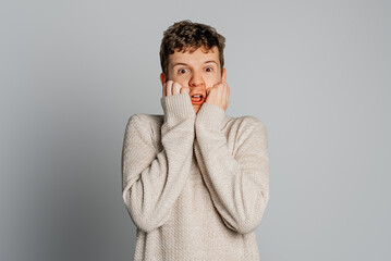 Portrait of handsome terrified young male being shocked to bottom of his heart, touches cheeks with both palms, notices something unexpected, standing over gray background