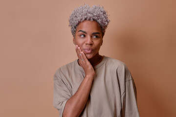 Young attractive African American woman shows that is shy, makes cute face with pouting lips and looks away due to uncomfortable situation holding cheeks posing in brown studio. People, lifestyle