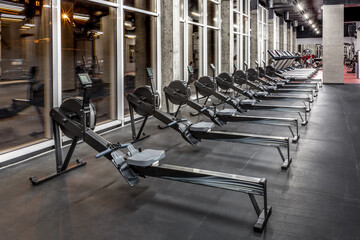 Lined modern rowing machines and treadmills by window in spacious, empty gym interior. Special...