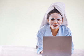 Portrait of young woman applying facial mask on her face during using laptop on bedroom