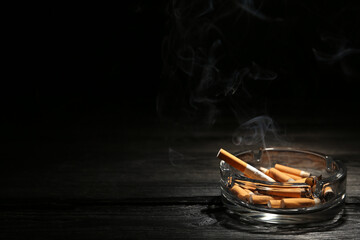 Smoldering cigarette in glass ashtray on dark wooden table against black background. Space for text