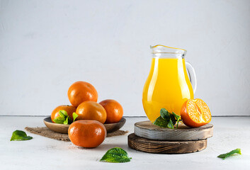 A glass jug of tangerine juice stands on round wooden stands. Whole tangerines on a wooden saucer in the background. Mint leaves on a light table