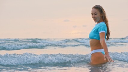SLOW MOTION GLOW MIST GOLD SUNSET CINEMATIC VIEW: Attractive woman standing on water sea. Girl stand in turquoise swimsuit. Freedom paradise holiday vacation summer beach, seaside landscape concept