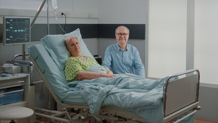 Portrait of pensioner with disease laying in bed and man giving assistance in hospital ward. Woman waiting on healthcare diagnosis and treatment while husband supports her at clinic.