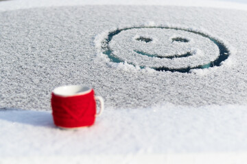 Fototapeta na wymiar A red mug with a hot drink on a snowy car trunk on a frosty sunny winter day. On the rear window of the car, a drawing of a smiling face. Selective focus. Close-up