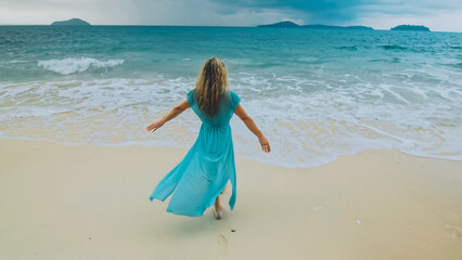 Charming woman with blonde curly long hair walk in beach, in stormy morning rain cloudy sea. Girl in turquoise swimsuit tunic. Concept resort outdoor relax summer vacation, travel. Dark dramatic view