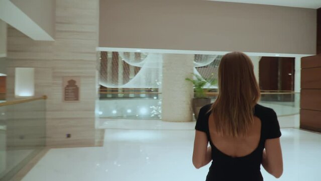 View from the back. A girl in a black dress with a plunging neckline exits the elevator. A woman steps out of an elevator in a luxury hotel.
