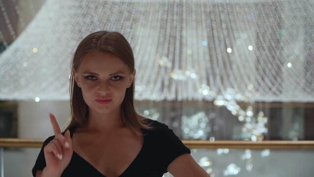 Caucasian girl gestures no sign. A beautiful European woman in a black dress waving her finger against the backdrop of a large crystal chandelier. The girl shows dislike.