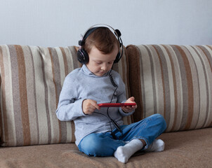 A 4-year-old boy in blue jeans and a gray turtleneck in headphones sits on the couch and looks...