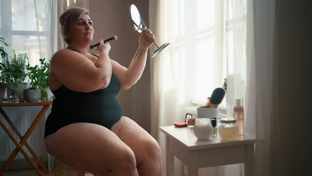 Overweight woman sitting and looking at mirror and applying make up at home, selfcare concept.