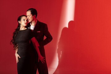 Creative portrait of tango dancers couple in dancing movement highlighted by a strip of light on red background. Handsome man in classic brown suit stands over a woman in elegant dress.